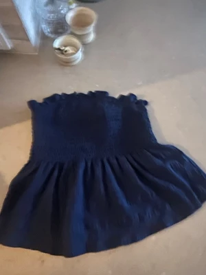 Mörkblå bandeau tubtopp - Snygg mörkblå bandeau tubtopp med smockad överdel och volangkant upptill. Toppen är axelbandslös och har en lätt utsvängd nederdel som ger en luftig känsla. Perfekt för varma dagar och enkel att styla till olika looks.