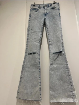 Ljusblå utsvängda jeans med slitningar - Säljer ett par ljusblå jeans med utsvängda ben och slitna detaljer vid knäna samt råa, fransiga benslut. Jeansen har klassisk femficksmodell och hög midja. Perfekta för en trendig och avslappnad look.