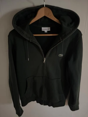 Mörkgrön hoodie från Lacoste - Snygg mörkgrön hoodie från Lacoste med dragkedja framtill och klassisk krokodil-logga på bröstet. Huva med dragsko och två stora fickor framtill. Perfekt för en chill och sportig stil.
