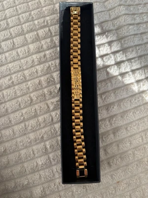 Rlx armband - Snyggt guldfärgat armband med bred länkdesign och rektangulära länkar. Armbandet har en stilren och modern look med en blank finish och ett dekorativt mönster på mitten. Perfekt accessoar för att lyfta din outfit.