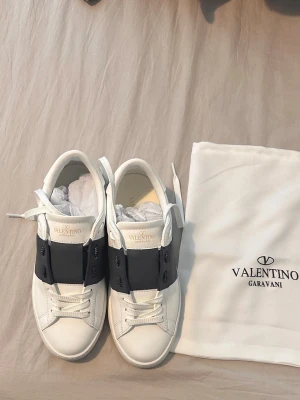 Valentino Garavani vita sneakers - Snygga vita sneakers från Valentino Garavani med bred svart läderdetalj över snörningen och klassisk logga på plösen. Skorna har en låg silhuett och är tillverkade i slätt läder. Medföljer original dustbag.