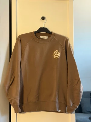 Brun sweatshirt från Les Deux - Snygg brun sweatshirt från Les Deux med broderad vit LD-logga på bröstet. Tröjan har rund hals, ribbade muddar och är tillverkad i mjuk bomull. Perfekt för en avslappnad och trendig stil.