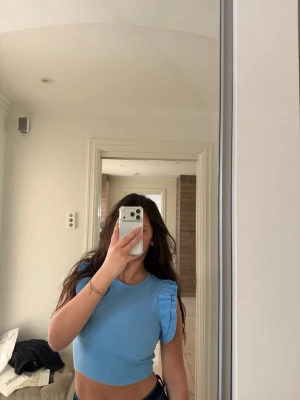 Ljusblå croppad topp med volang - Trendig ljusblå croppad topp med volangdetaljer på axlarna och ribbad struktur. Perfekt för dig som vill ha en söt och stilren look. Toppen är ärmlös och har en tight passform som framhäver figuren.