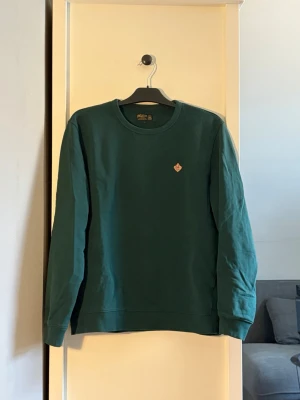 Grön sweatshirt från Morris  - Mörkgrön sweatshirt från Morris med rund halsringning och diskret logotyp på bröstet. Tröjan har långa ärmar och ribbade muddar vid ärmslut och nederkant. Perfekt för en avslappnad och stilren look.