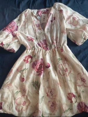 Blommig kortklänning med puffärm - Söt kortklänning från HM i ljusbeige med blommigt mönster i rosa och grönt. Klänningen har v-ringning, puffiga kortärmar och markerad midja. Perfekt för varma dagar och somriga tillfällen.