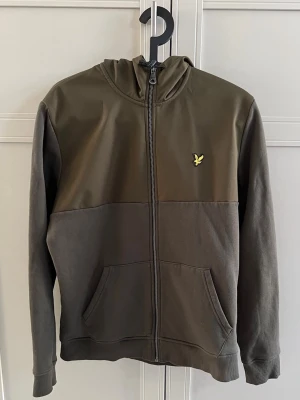 Olivgrön hoodie från Lyle & Scott - Snygg olivgrön hoodie från Lyle & Scott med dragkedja framtill och klassisk gul logga på bröstet. Hoodien har kängurufickor, ribbade muddar och en mjuk bomullsblandning. Perfekt för en avslappnad streetwear-look.