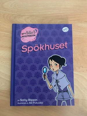 Spökhuset - Ett Billie B-mysterium - En spännande och färgglad barnbok med lila hård pärm, titeln 'Spökhuset' och illustration av en ung detektiv. Boken är skriven av Sally Rippin och illustrerad av Aki Fukuoka. Perfekt för dig som gillar mysterier och äventyr.