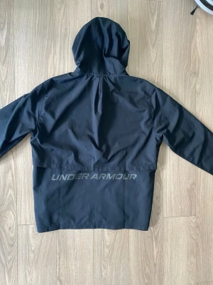 Svart jacka från Under Armour - Svart hoodie från Under Armour med dragkedja framtill och två sidofickor. Jackan har en stor huva och en diskret logga på ryggen. Tillverkad i ett lätt och smidigt syntetmaterial som känns sportigt och modernt. Knappt använd 