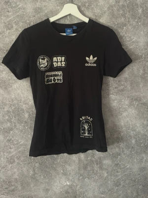 Adidas t-shirt med tryck - Säljer en svart t-shirt från Adidas med flera vita tryck och loggor på framsidan. T-shirten har rund hals, korta ärmar och är i en klassisk passform. Perfekt för dig som gillar streetwear och sportig stil.