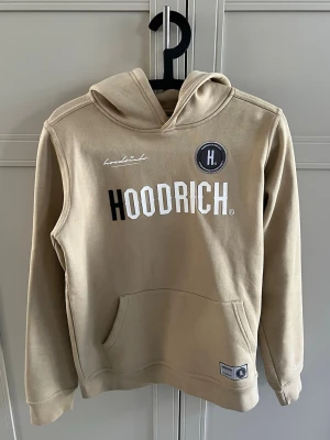 Beige hoodie från Hoodrich - Snygg beige hoodie från Hoodrich med stor logga framtill och tryck på ryggen. Hoodien har huva, känguruficka och ribbade muddar. Perfekt för en avslappnad streetwear-look. Materialet är mjukt och skönt.