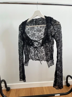 Svart spetsblus med snörning - Supersnygg svart blus i transparent spets med långa ärmar och snörning framtill. Blusen har volangdetaljer vid halsringningen och är perfekt för dig som vill sticka ut. Den är croppad och har ett romantiskt mönster i spets.