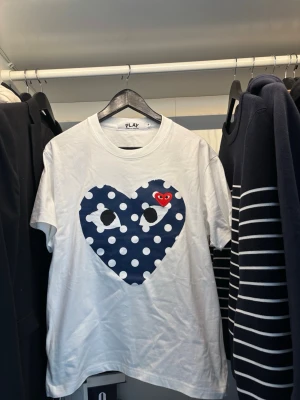 Vit Comme des Garçons Play t-shirt - Vit t-shirt från Comme des Garçons Play med stort blått hjärta med vita prickar och ögon på bröstet. Liten röd hjärtpatch med ögon broderad ovanpå. Klassisk rund hals och korta ärmar. Perfekt statement-plagg för en trendig look.