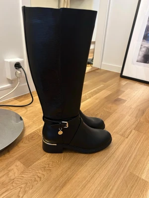 Svarta höga stövlar med gulddetalj - Snygga svarta knähöga stövlar med guldfärgad detalj vid hälen och dekorativt spänne med hängande berlock. Stövlarna har en slät framsida och en baksida med diskret mönster, samt dragkedja på insidan. Aldrig använda 