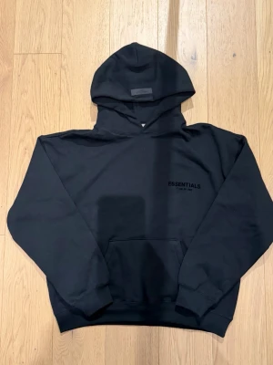 Svart Essentials Fear of God hoodie - Svart hoodie från Fear of God Essentials med diskret logga på bröstet och stor logga på ryggen. Klassisk känguruficka framtill, ribbade muddar och huva. Perfekt för en avslappnad streetwear-look.