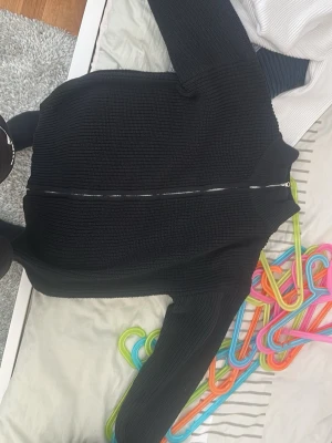 Svart stickad ziptröja - Svart stickad ziptröja med grov ribbstickning och hel dragkedja framtill. Tröjan har hög krage och långa ärmar, perfekt för lager-på-lager. Enkel och stilren design som passar till det mesta.