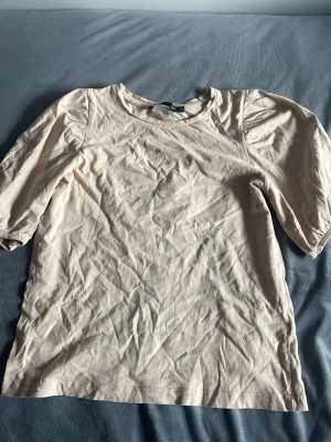 Beige topp med puffärm från 157 - Snygg beige topp från 157 med puffiga ärmar och rund halsringning. Tillverkad i mjuk bomull och har en enkel, stilren design som passar perfekt till jeans eller kjol.
