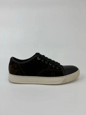 Bruna Lanvin sneakers med mockadetaljer - Stilrena bruna sneakers med ovandel i mocka och tåparti i slätt läder. Skorna har svarta skosnören, silverfärgade öljetter och en vit, klassisk gummisula. Perfekta för dig som gillar en clean och tidlös look.