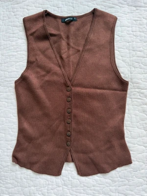 Brun stickad väst med knappar - Snygg brun stickad väst från Gina Tricot med v-ringning och knappar framtill. Inga fler bilder då det är packat i flyttlåda.