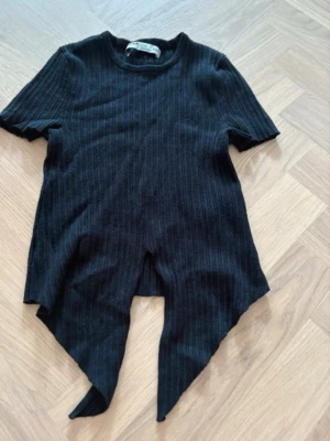 Svart ribbad topp från FB Sister - Cool svart ribbad topp från FB Sister med korta ärmar och unik asymmetrisk nederkant. Toppen har rund halsringning och är tillverkad i ett stretchigt material som sitter snyggt på kroppen. Perfekt för dig som gillar edgy och trendiga plagg.