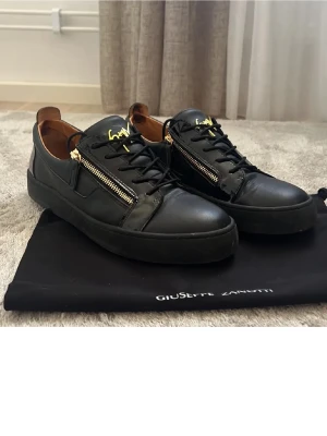 Svarta Giuseppe Zanotti  - Snygga svarta sneakers från Giuseppe Zanotti i läder med guldfärgade dragkedjor på sidorna och guldfärgad logga på plösen. Skorna har svarta snören och en chunky svart sula. Perfekta för dig som gillar en lyxig och edgy stil.