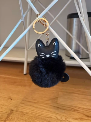 Nyckelring med svart katt och fluffboll - Söt nyckelring med svart kattmotiv i konstläder och en fluffig svart boll. Har guldfärgad metallring och karbinhake. Perfekt att hänga på väskan eller nycklarna för en gullig touch.
