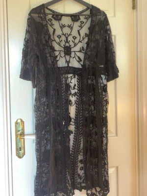 Svart transparent strandkimono - Säljer en svart transparent strandkimono med vackra broderade blommor och korta ärmar. Kimono-modellen har öppen front och är perfekt att slänga över bikinin på stranden. Materialet är tunt mesh med detaljerade broderier för en bohemisk look.
