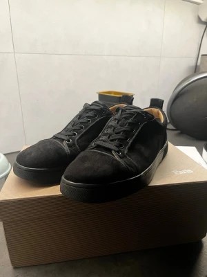 Svarta sneakers från Christian Louboutin - Stilrena svarta sneakers i mocka från Christian Louboutin med diskret logga broderad på hälen. Skorna har svarta skosnören, svart sula och en låg siluett. Insidan är fodrad i beige läder för extra komfort och lyxig känsla.