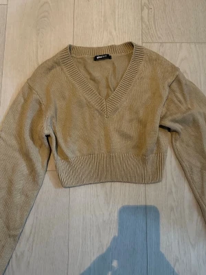 Beige cropped stickad tröja Gina Tricot - Trendig beige stickad tröja från Gina Tricot med croppad passform och djup V-ringning. Långa ärmar och bred ribbad mudd vid midjan och halsen. Perfekt för lager-på-lager och enkel att styla till olika outfits.