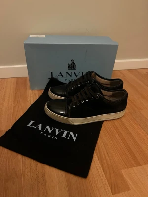 Svarta sneakers från Lanvin - Stilrena svarta sneakers från Lanvin med vit sula och lackad tå. Skorna har svarta skosnören och är tillverkade i en mix av mocka och läder. Kommer med originalkartong, dustbag och extra snören för extra lyxig känsla. Är i väldigt bra skick!