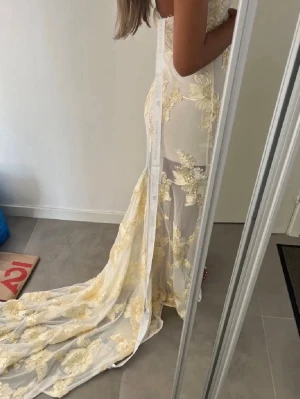 Balklänning - Säljer en elegant axelbandslös klänning med långt släp och gulfärgade blommiga broderier. Klänningen har figurnära passform och transparenta partier som ger en lyxig känsla. Perfekt för bal eller speciella tillfällen.