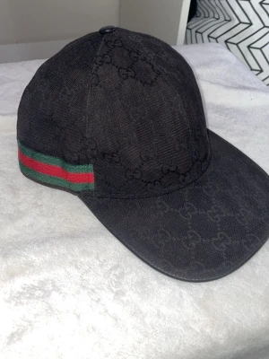 Svart Gucci keps med GG-mönster - Säljer en svart keps från Gucci med det klassiska GG-monogrammet över hela kepsen. På sidan finns en grön och röd rand som ger en ikonisk touch. Kepsen har böjd skärm och justerbar rem bak. Materialet är canvas med detaljer i bomull. Billigare pris vid smidig och snabb affär 
