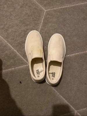 Beige slip-on sneakers från 157 - Stilrena slip-on sneakers i beige canvas från 157. Skorna har en klassisk låg profil med vit sula och elastiska paneler på sidorna för enkel påtagning. Perfekta för en avslappnad och trendig look.
