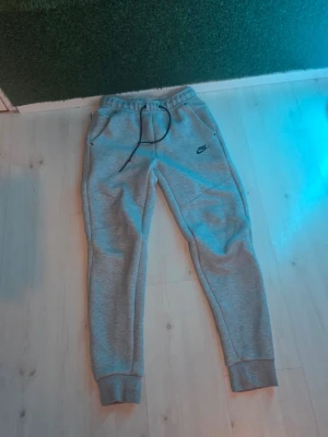 Gråa mjukisbyxor från Nike - Snygga gråa mjukisbyxor från Nike med svart logga på vänster ben. Byxorna har resår och dragsko i midjan, två sidofickor och muddar vid bensluten. Perfekta för chill eller träning, tillverkade i mjukt och bekvämt material.