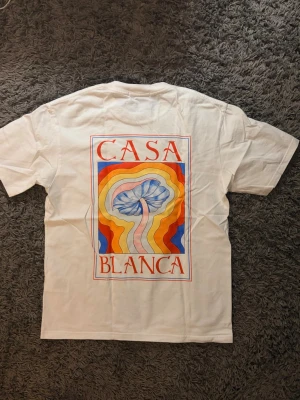 Vit t-shirt från Casablanca med tryck - Vit t-shirt från Casablanca med färgstarkt grafiskt tryck på både bröstet och ryggen. Trycket har vågiga linjer i orange, blått, gult och rött samt texten 'Casa Blanca'. Klassisk passform och rund hals. Perfekt för dig som gillar statement-plagg.