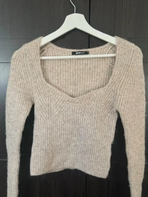 Beige stickad tröja Gina Tricot - Snygg beige stickad tröja från Gina Tricot med ribbstickat mönster och djup, fyrkantig urringning. Tröjan har långa ärmar och är tillverkad i ett mjukt, mysigt material som passar perfekt till kalla dagar.