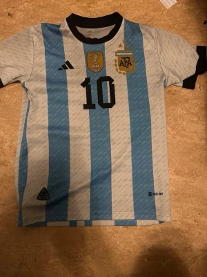 Argentina Messi matchtröja Adidas - Säljer en Argentina fotbollströja från Adidas med Messis namn och nummer 10. Tröjan har blå och vita ränder, svarta detaljer vid krage och ärmslut samt officiella tryck och märken från FIFA och AFA. Perfekt för dig som älskar fotboll och Messi.