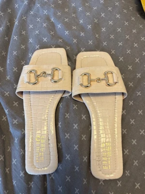 Beige sandaler med krokopräglat mönster - Stilrena beige sandaler från Ego med fyrkantig tå och krokopräglat mönster. Dekorativa silvriga metalldetaljer framtill ger en lyxig känsla. Tillverkade i syntetmaterial med en bred rem över foten. Perfekta för en trendig och chic look.