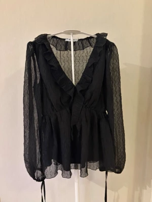 Svart volangblus med transparenta ärmar - Säljer en svart blus från NA-KD i storlek 32. Blusen har volanger framtill och transparenta armar. Har aldrig kommit till användning. 