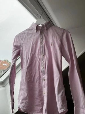 Ljusrosa skjorta från Ralph Lauren - Snygg ljusrosa skjorta från Ralph Lauren med klassisk button-down krage och broderad logga på bröstet. Skjortan har långa ärmar och vita knappar framtill. Perfekt för en stilren och avslappnad look. Skriv vid frågor!