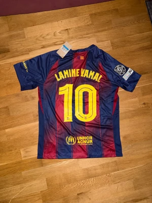 FC Barcelona matchtröja Lamine Yamal - Säljer en officiell FC Barcelona matchtröja från Nike med Lamine Yamal och nummer 10 på ryggen. Tröjan har blå och vinröda ränder, gula detaljer och klubbmärke samt sponsorloggor på bröstet. Tillverkad i lätt och ventilerande material, perfekt för fotbollsträning.