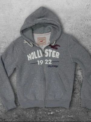Grå Hollister hoodie med dragkedja - En fet zip hoodie från hollister i storlek M även fast det står XL. Färg grå med vitt tryck. Bra skick! Vid minsta lilla fråga skriv🤙