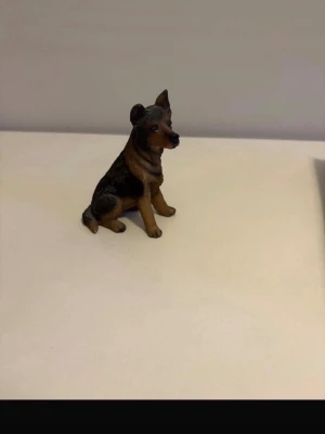 Hundfigur i porslin, brun och svart - Säljer en liten hundfigur i porslin som föreställer en sittande hund med bruna och svarta detaljer. Perfekt som dekoration eller samlarobjekt. Realistisk design med fina detaljer på pälsen och ansiktet.