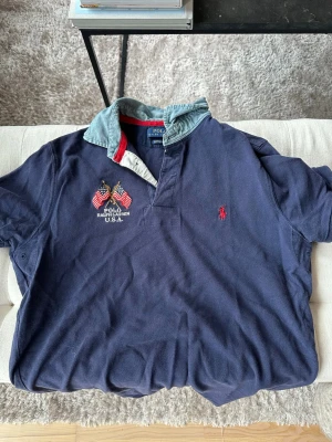 Blå pikétröja från Polo Ralph Lauren - Mörkblå pikétröja från Polo Ralph Lauren med broderade amerikanska flaggor och logga på bröstet. Kragen är i ljus denim med röd insida. Klassisk passform och korta ärmar, perfekt för en avslappnad stil.