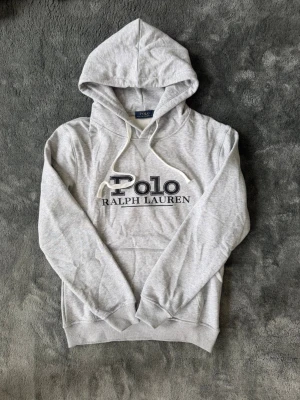 Grå hoodie från Polo Ralph Lauren - Snygg ljusgrå hoodie från Polo Ralph Lauren med stor svart logga framtill. Klassisk modell med huva och vita dragsnören, samt magficka. Perfekt för en avslappnad och trendig stil.