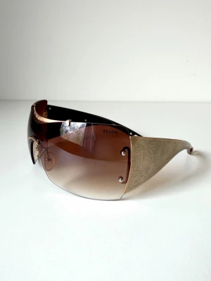Dior bruna shield-solglasögon - Snygga shield-solglasögon från Dior med stora, bruna tonade glas och guldfärgade sidor med ingraverat mönster. Har tyvärr inga intyg på att de är äkta, därav pris. Priset går att diskutera 🤍
