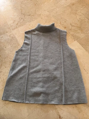 Grå ärmlös stickad poloväst - Snygg grå stickad väst med polokrage och ärmlös design. Västen har en rak och något utsvängd passform med ribbstickad krage. Perfekt att styla över en skjorta eller långärmad tröja för en trendig lager-på-lager-look.
