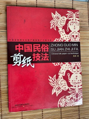 Chinese folk paper-cut technique bok - En bok med röd framsida och vita detaljer som handlar om kinesisk pappersklippning. Innehåller illustrationer och instruktioner på kinesiska och engelska. Perfekt för dig som vill lära dig traditionell kinesisk konst och hantverk.
