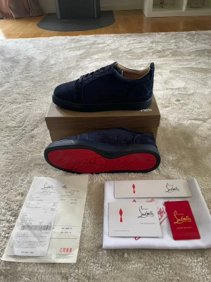 Mörkblå Christian Louboutin sneakers - Snygga mörkblå sneakers från Christian Louboutin i mocka med klassisk röd sula och diskret logga. Skorna har lågt skaft, svarta skosnören och exklusiva detaljer. Perfekta för dig som vill sticka ut med stilrena och lyxiga sneakers. Skorna är helt nya och allt orginal följer med.