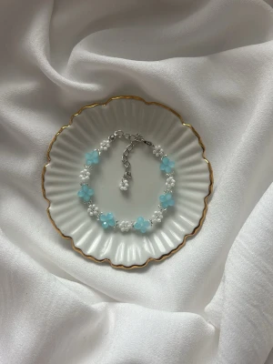 Blått och vitt pärlarmband med blommor - Sött armband med små vita pärlor och ljusblå blommor. Armbandet har justerbar silverfärgad kedja och är perfekt för dig som gillar en lekfull och trendig stil. Tillgänglig i andra färger