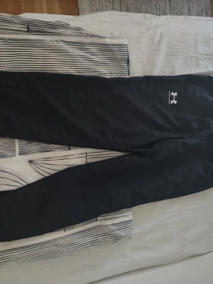 Svarta mjukisbyxor från Under Armour - Svarta mjukisbyxor från Under Armour med smal passform och diskret logga på ena låret. Byxorna har elastisk midja och är tillverkade i ett mjukt och stretchigt material som passar perfekt för chill eller träning. Perfekt för dig som vill se bra ut i sommar. 